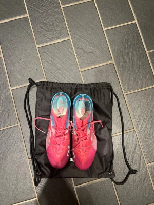 Puma rosa/blå fotbollsskor - Snygga fotbollsskor från Puma i en fräsch rosa och blå färgkombination. Skorna har snörning, låg profil och är tillverkade i ett lätt syntetmaterial för bästa känsla på planen. Perfekta för dig som vill sticka ut och ha bra grepp på gräs.