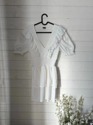 Vit volangklänning från Gina Tricot - Supersöt vit kortklänning från Gina Tricot med v-ringning, volangdetaljer och puffärmar. Klänningen har smock i midjan och är tillverkad i ett lätt, strukturerat tyg som känns perfekt för varma dagar.