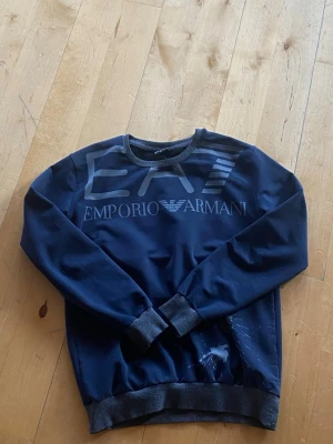 Mörkblå sweatshirt från Emporio Armani - Mörkblå sweatshirt från Emporio Armani med stor EA7-logga och text framtill. Tröjan har används i ungefär 1,5 år, fungerar fortfarande som ny. 