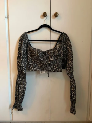 Blommig croppad blus med snörning - Trendig svart blommig blus med vita små blommor, croppad passform och långa puffärmar med smock vid ärmslut. Söt snörning framtill och bred urringning. Perfekt för dig som vill ha en romantisk och cool look.