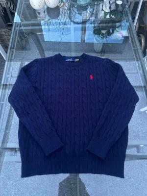 Ralph Lauren tröja - Mörkblå kabelstickad tröja från Polo Ralph Lauren med klassisk röd broderad logga på bröstet. Tröjan har rund halsringning, ribbade muddar och nederkant. Perfekt för dig som gillar tidlös och stilren design.