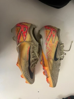 Nike Mercurial fotbollsskor beige/orange - Säljer ett par Nike Mercurial fotbollsskor i beige med orange och röda detaljer samt svart Nike-logga. Skorna har snörning och en slitstark ovandel i syntetmaterial. Sulan är orange med dobbar för bra grepp på planen. Perfekta för dig som vill sticka ut på fotbollsplanen.