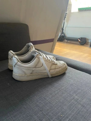 Vita Nike Air Force 1 låga sneakers - Klassiska vita Nike Air Force 1 sneakers i lågt utförande. Skorna har en stilren design med vit ovandel i läder, vita skosnören och den ikoniska swoosh-loggan på sidan. Perfekta för dig som gillar en clean och tidlös look.