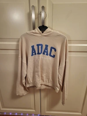 Beige hoodie med blå ADAC-tryck - Säljer en beige hoodie med blått ADAC-tryck på bröstet och texten 'Local Adventure Camp' under. Hoodien har huva och avslappnad passform med resår i nederkanten. Perfekt för en chill och sportig look.