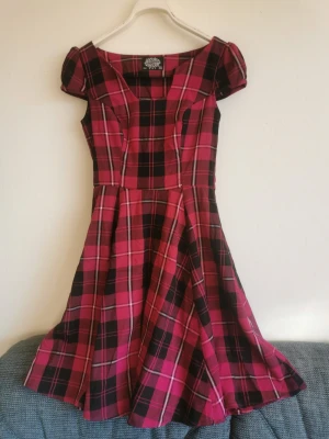 Rosa/svart rutig klänning  - Knälång rosa/svart rutig klänning i flanelltyg. Känns mer som storlek 34 eller 36 än 38. Har köpt ett svart sidenband som jag hade tänkt fästa som en rosett över urtaget i halsringningen, kan skicka med det om intresse finns. 