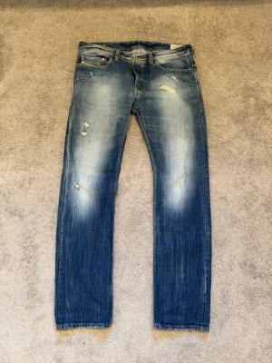 Blå slitna jeans från Diesel - Säljer ett par blå jeans från Diesel med slitningar och hål på låret och knät. Jeansen har en klassisk femficksmodell, raka ben och snyggt tvättad finish med ljusare partier. Perfekta för en avslappnad och trendig look.