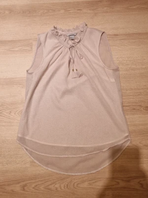 Beige ärmlös blus med volang - Säljer en ljusbeige ärmlös blus från H&M med volangdetalj vid halsen och knytband framtill. Blusen är tillverkad i ett lätt och luftigt polyestertyg som ger en snygg flowy känsla. Perfekt till sommaren och enkel att styla.