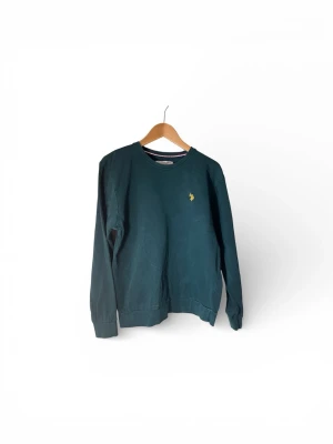 Mörkgrön sweatshirt från U.S. Polo Assn. - Mörkgrön sweatshirt från U.S. Polo Assn. med rund halsringning och gul broderad logga på bröstet. Tröjan har långa ärmar och ribbade muddar vid ärmslut och nederkant. Perfekt för en avslappnad och stilren look.