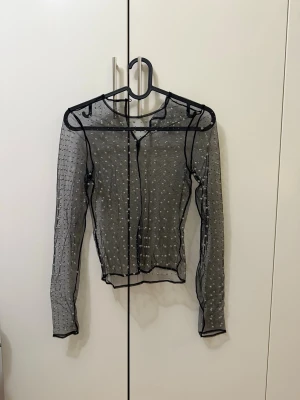 Svart meshblus med prickar - Snygg transparent svart meshblus med små vita prickar och markerade svarta sömmar. Blusen har lång ärm och rund halsringning. Perfekt att styla lager på lager för en trendig look.