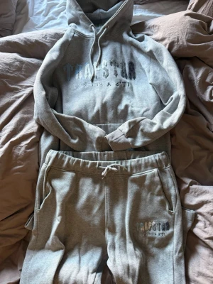 Grått set från Trapstar med tryck - Säljer ett grått mjukisset från Trapstar med huva och dragsko på tröjan samt matchande byxor med resår och snörning i midjan. Båda delarna har trycket 'Trapstar It's a Secret' framtill. Perfekt för en chill och avslappnad stil. Storlek L men passar M