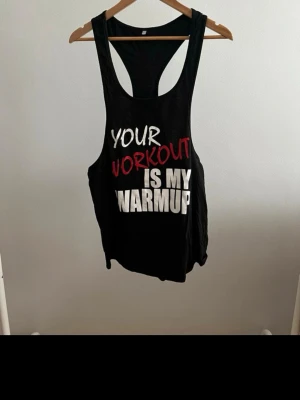 Svart träningslinne med tryck - Säljer ett svart träningslinne med brottarrygg och stort tryck framtill: 'YOUR WORKOUT IS MY WARMUP' i vitt och rött. Perfekt för gymmet eller löpning. Linnet är gjort i ett mjukt och lätt material som andas bra.