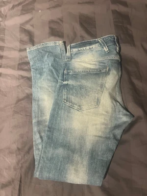 Blå slitna jeans med tvättade detaljer - Säljer ett par blå jeans med snygga slitningar och snygg tvätt. Ett par replay anbass jeans med normal midja och slim i passform. Jeansen har en ljusare tvätt framtill och baktill för en cool, avslappnad look.