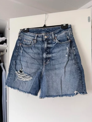 Mörkblå jeansshorts med slitningar från H&M, storlek S - Säljer ett par coola blå jeansshorts från H&M med slitningar och fransiga kanter. Shortsen har en hög midja och klassisk femficksdesign. Perfekta för en avslappnad stil. Använda ca 5 gånger. 