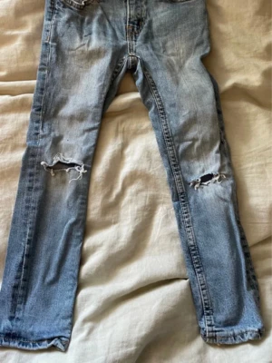 Ljusblå slitna jeans med hål - Säljer ett par ljusblå jeans med raka ben och slitna detaljer på båda knäna. Pojkbyxor i storlek 120. Jeansen har klassisk femficksmodell och är tillverkade i denim. 