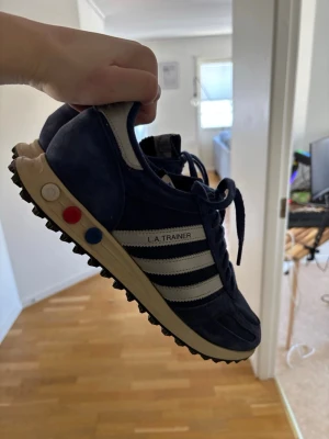 Adidas L.A. Trainer blå sneakers - Klassiska Adidas L.A. Trainer sneakers i marinblå mocka med vita detaljer och tre ränder på sidan. Skorna har en retrodesign med färgade pluggar i sulan och grovt mönstrad yttersula för bra grepp. Perfekta för dig som gillar old school-stil.