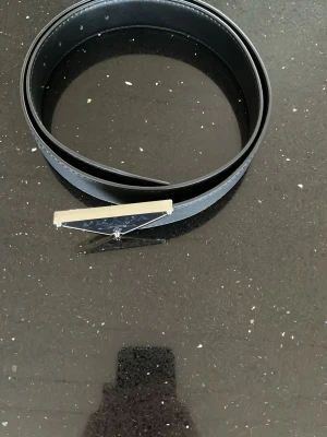 Svart skärp från hm - Snyggt svart skärp från hm  med ett stilrent trekantigt metallspänne där loggan syns tydligt. Skärpet är gjort i slätt skinn och har en klassisk, minimalistisk design som passar till många olika looks.