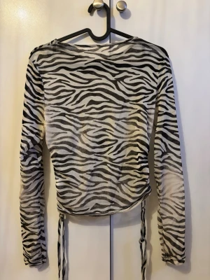 Zebramönstrad mesh topp med snörning - Säljer en långärmad zebramönstrad mesh topp i svart och vitt. Toppen har snörning i sidorna för en justerbar passform och är tillverkad i ett tunt, genomskinligt syntetmaterial. Perfekt för dig som vill sticka ut med ett djurmönster! Det finns lite slitningar i armveck och armhåla pga användning.