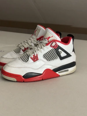 Air Jordan 4 Retro Fire Red - Säljer ett par Air Jordan 4 Retro Fire Red sneakers med vit ovandel, röda och svarta detaljer samt meshpaneler på sidorna. Klassisk Jumpman-logga på plös och sula. Skorna har en chunky sula och ikoniska plastdetaljer vid snörningen. Skriv vid intresse!