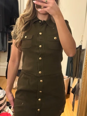 Olivgrön kortärmad skjortklänning - Snygg olivgrön sett med korta ärmar och guldknappar framtill. Klänningen har två bröstfickor och markerad midja för en stilren look. Perfekt för dig som gillar klassiska detaljer och enkel design. Kjol: S Topp: XXS