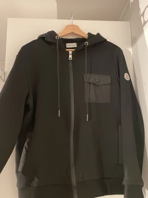 Svart hoodie från Moncler med dragkedja - (Alldrig använd) Snygg svart hoodie från Moncler med dragkedja framtill, huva med dragsko och en bröstficka med lock. Ikonisk Moncler-logga på ärmen och diskreta sidofickor. Perfekt för dig som vill ha en stilren och exklusiv look. (Byxor finns också med att köpa för endast 200kr)