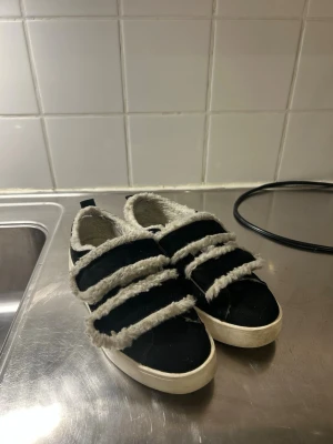 Svarta sneakers med fuskpälsdetaljer - Svarta sneakers med tre breda kardborreband och vit sula. Skorna har mjuk fuskpäls längs kanterna och insidan, vilket ger en mysig känsla. Perfekta för dig som vill ha något unikt och bekvämt. Passar till en avslappnad stil.