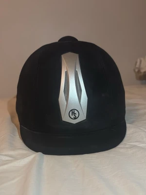 Svart ridhjälm från HS - Säljer en svart ridhjälm från HS med en snygg silverfärgad detalj framtill. Hjälmen har rundad form, justerbar rem och ventilationsöppningar. Passar perfekt för barn, men även vuxna.