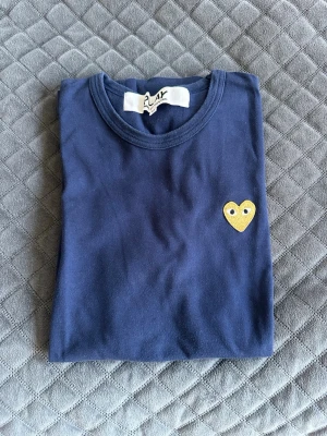 Blå Comme des Garçons PLAY t-shirt - Säljer en mörkblå t-shirt från Comme des Garçons PLAY med ett gult hjärtat broderat på bröstet. Klassisk rund halsringning och mjuk bomullskvalitet. Perfekt för dig som gillar stilrena plagg.  Perfekt inför sommaren och är i väldigt bra skick!! Skriv om ni är intresserade om mer info eller bilder. Kan dock tyvärr inte vissa passform för den är för liten.