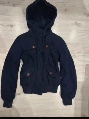 Marinblå vår/höst jacka - Snygg marinblå jacka med beige quiltat innerfoder. Jackan har ribbade muddar, dragkedja framtill och fyra fickor med dekorativa knappar. Perfekt för dig som vill ha en stilren och bekväm jacka med streetkänsla.