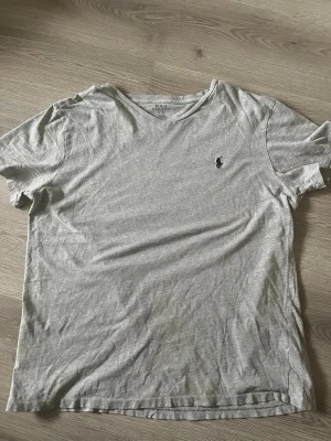 Grå t-shirt från Polo Ralph Lauren - Klassisk grå t-shirt från Polo Ralph Lauren med korta ärmar och rund hals. T-shirten har en liten mörkblå logga broderad på bröstet och är tillverkad i mjuk bomull. Perfekt basplagg för en avslappnad stil. Storlek L
