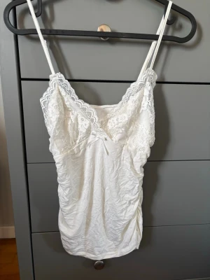 Vit spetsdetaljerad linnetopp - Söt vit linnetopp med tunna axelband och v-ringad spetskant upptill. Toppen har en liten rosett framtill och är gjord i ett mjukt, stretchigt material som känns bekvämt mot huden. Perfekt att styla lager på lager eller bära som den är.