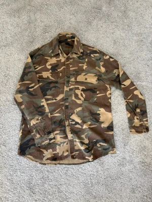 Camo overshirt med bröstfickor - Snygg overshirt i klassiskt camouflagemönster med två stora bröstfickor och knäppning framtill. Skjortan har en rak passform, krage och är tillverkad i ett slitstarkt bomullstyg. Perfekt för lager på lager och streetwear-stil. Köpt i USA när jag var där men står inget märke i den! Skickar den självklart inom 24h och efter köp, hör av dig vid frågor och fler bilder! 