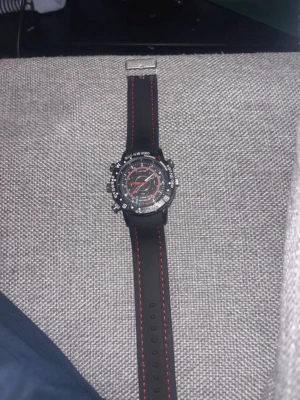 Spionklocka- Svart sportklocka med röd detalj - - Säljer en svart sportig klocka med silikonarmband och röda sömmar. Urtavlan är svart med vita och röda detaljer samt flera knappar runt boetten. Klockan har en robust design och tydliga markeringar för kompassriktningar på ringen.