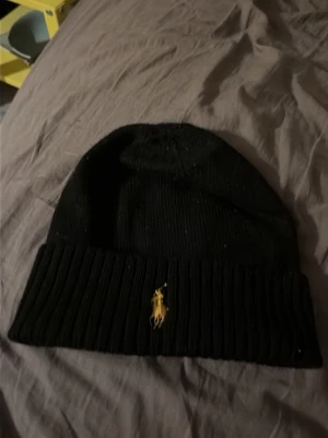 Svart ribbstickad mössa från Polo Ralph Lauren - Säljer en svart ribbstickad mössa från Polo Ralph Lauren med den klassiska gula loggan broderad framtill. Mössan har uppvikt kant och är tillverkad i mjukt material som håller dig varm under kalla dagar. Perfekt accessoar för en stilren look.