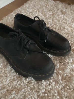 Svarta loafers från Dr. Martens - Klassiska svarta loafers från Dr. Martens i äkta läder med rund tå och grov sula. Skorna har snörning och den ikoniska gula sömmen runt sulan. Perfekta för dig som gillar en tidlös och edgy stil.