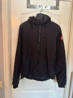 Svart vindjacka från Canada Goose - Snygg svart vindjacka från Canada Goose med huva och dragkedja framtill. Jackan har en bröstficka med dragkedja och två sidofickor. Klassisk logotyp på ärmen och justerbar nederkant. Perfekt för blåsiga dagar och streetwear-stil.