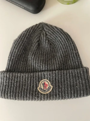 Grå ribbstickad mössa från Moncler - Säljer en snygg grå ribbstickad mössa från Moncler med uppvikt kant och klassisk Moncler-logga framtill. Mössan är tillverkad i mjukt material som håller dig varm och passar perfekt till höst och vinter. Enkel att matcha med olika outfits. 
