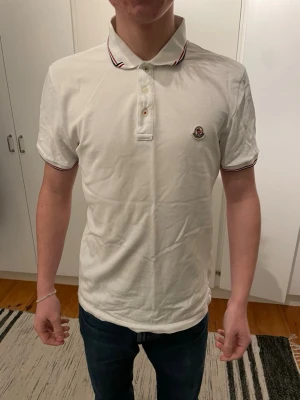 Vit Moncler pikétröja med röd detalj - Klassisk vit pikétröja från Moncler med korta ärmar och knappslå framtill. Tröjan har Moncler-logga på bröstet och röda samt svarta ränder på krage och ärmslut för en sportig touch. Tillverkad i bomull och har en normal passform.