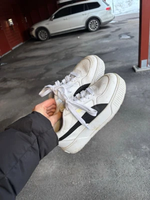 Vita Puma sneakers med svart detalj - Säljer ett par fräscha vita Puma sneakers med svart sidodetalj och guldfärgade loggor. Skorna har tjock sula, klassisk låg modell och är tillverkade i läder med perforerade detaljer framtill. Perfekta för dig som gillar stilrena och sportiga sneakers. Storlek 37 använda två gånger 