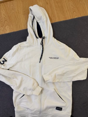 Vit hoodie från Race Marine - Snygg vit hoodie från Race Marine med dragkedja framtill och huva med snörning. Broderad logga på bröstet och siffran 3 på ärmen. Två fickor framtill och mörkblå detaljer vid fickor och huva. Perfekt för en avslappnad och sportig stil.