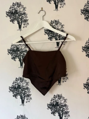 Brun bandana-linnetopp i satin - Snygg brun linnetopp i bandana-stil med smala axelband och spetsig nederkant. Toppen är gjord i ett glansigt satinliknande material och har smockad rygg för extra komfort. Perfekt för dig som vill ha en trendig och stilren look.