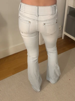 Ljusblå bootcut jeans med låg midja - Säljer ett par ljusblå bootcut jeans med låg midja och dubbla knappar framtill. Jeansen har klassiska bakfickor och framfickor med detaljer i silver. Perfekta för en trendig och avslappnad stil. 