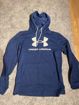 Blå hoodie från Under Armour - Snygg blå hoodie från Under Armour med stor vit logga och text på bröstet. Hoodien har huva med dragsko och en stor magficka framtill. Perfekt för en avslappnad och sportig stil.