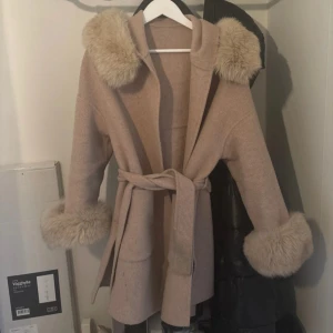 Beige kappa med ältapäls och huva - Snygg beige kappa med huva och breda äktapälsdetaljer på både ärmslut och huva. Kappan har knytskärp i midjan och en rak, avslappnad passform. Perfekt för dig som vill ha en trendig och mysig look under kallare dagar.