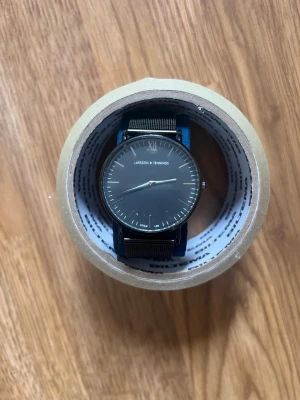 Svart klocka från Larsson & Jennings - Stilren svart klocka från Larsson & Jennings med rund urtavla och mesh-armband i metall. Klockan har minimalistisk design med tunna visare och diskreta markeringar. Perfekt accessoar för en modern look.