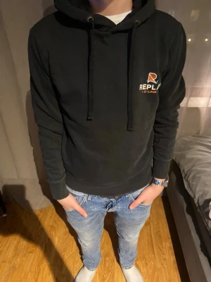 Svart hoodie från Replay - Snygg svart hoodie från Replay med justerbar huva och dragsnören. Liten logga i vitt och orange på bröstet. Klassisk passform och mjukt material som känns skönt mot huden. Perfekt till jeans eller joggers för en avslappnad look.