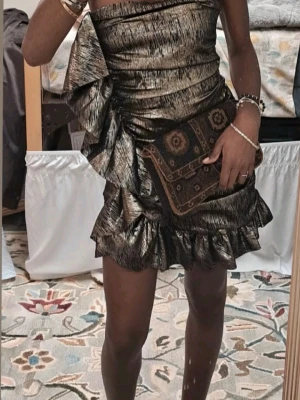 Glittrig svart och guld kortklänning - Snygg axelbandslös kortklänning i svart och guld med glittrig finish från Zara. Klänningen har volangdetaljer och en stor dekorativ rosett på ena sidan. Perfekt för dig som vill sticka ut med en unik look.