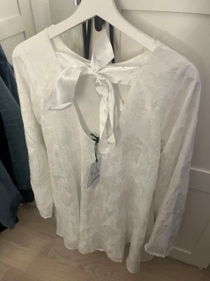 Vit blus med rosett och blommönster - Superfin vit blus med subtilt blommönster och lång ärm. Blusen har en snygg öppen rygg med bred satinrosett som knyts i nacken. Tunt, lätt tyg som ger en luftig känsla och elegant look.