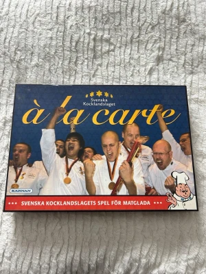 À la carte - Svenska Kocklandslaget - Säljer boken 'À la carte' från Svenska Kocklandslaget, utgiven av Kärnan. Boken har ett blått omslag med guldtext och bilder på kockar. Innehåller spel och matglada utmaningar, perfekt för dig som gillar mat och tävling.