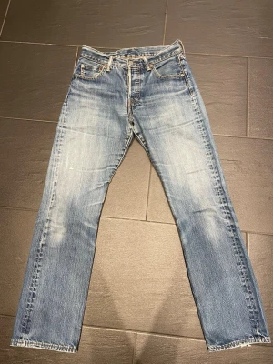 Levi's blå raka jeans - Säljer ett par klassiska blå jeans från Levi's med raka ben och snyggt slitna detaljer. Jeansen har fem fickor, dragkedja och knappstängning samt den ikoniska Levi's-lappen på bakfickan. Perfekt för dig som gillar tidlös denimstil. Trasig lapp men inget som påverkar jeansen, ställ frågor vid fundering!