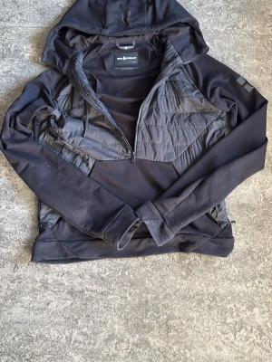 Svart cardigan från sail racing - Svart jacka från sail racing med huva och quiltade partier framtill. Jackan har lång ärm, dragkedja framtill och är tillverkad i en mix av syntetmaterial och polyester. Perfekt för dig som gillar en sportig och stilren look.